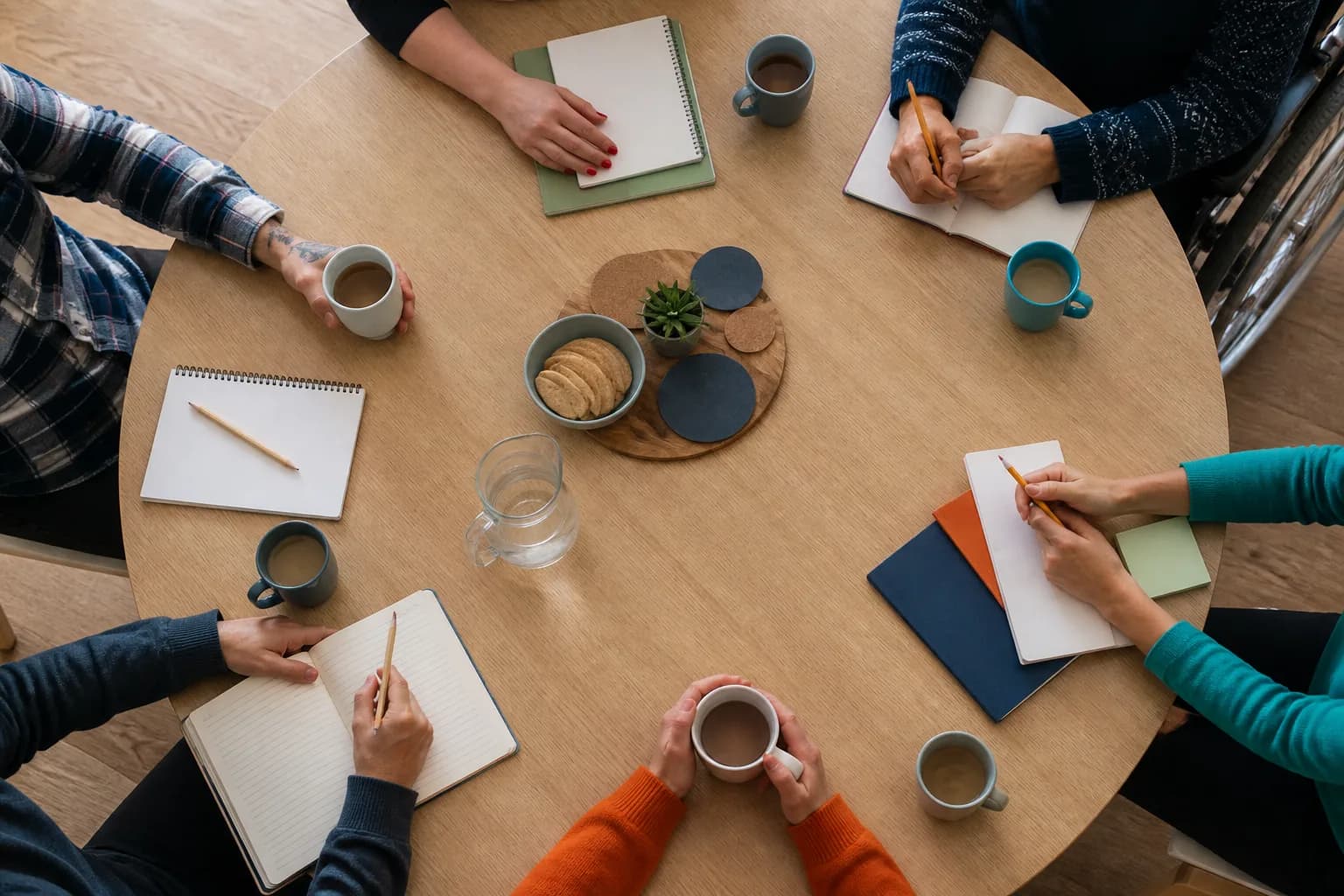 Hands of peer-support participants around a shared table with notebooks, refreshments and accessible seating